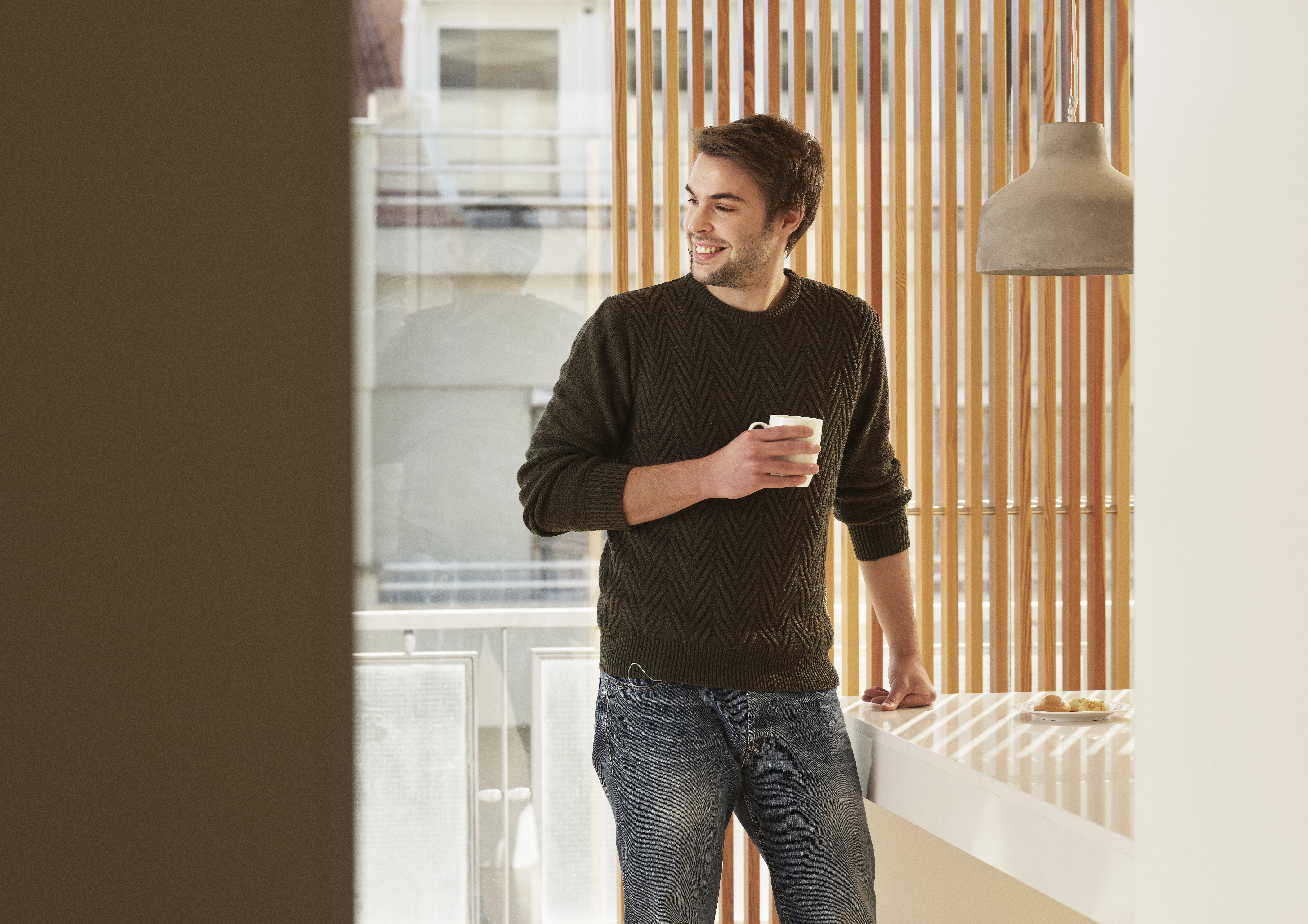 Ein dunkelhaariger Mann steht vor einem Fenster und hält eine Tasse in der Hand. 